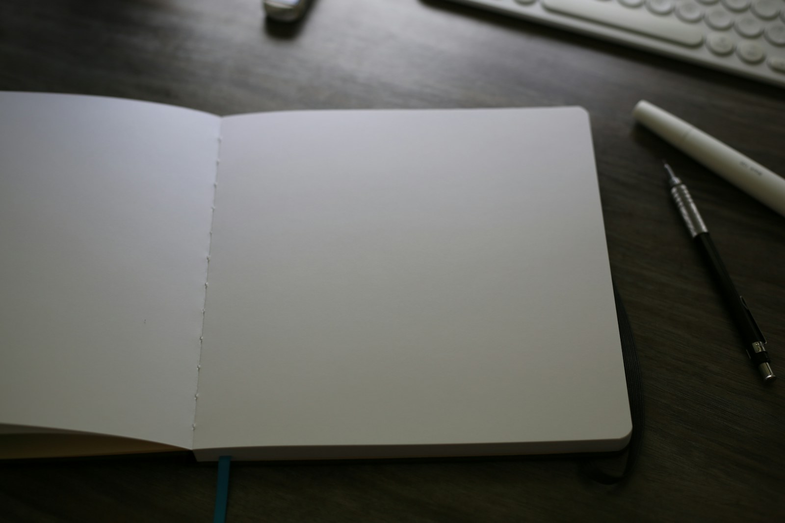 An open blank notebook on a dark desk beside a keyboard, pencil and marker.