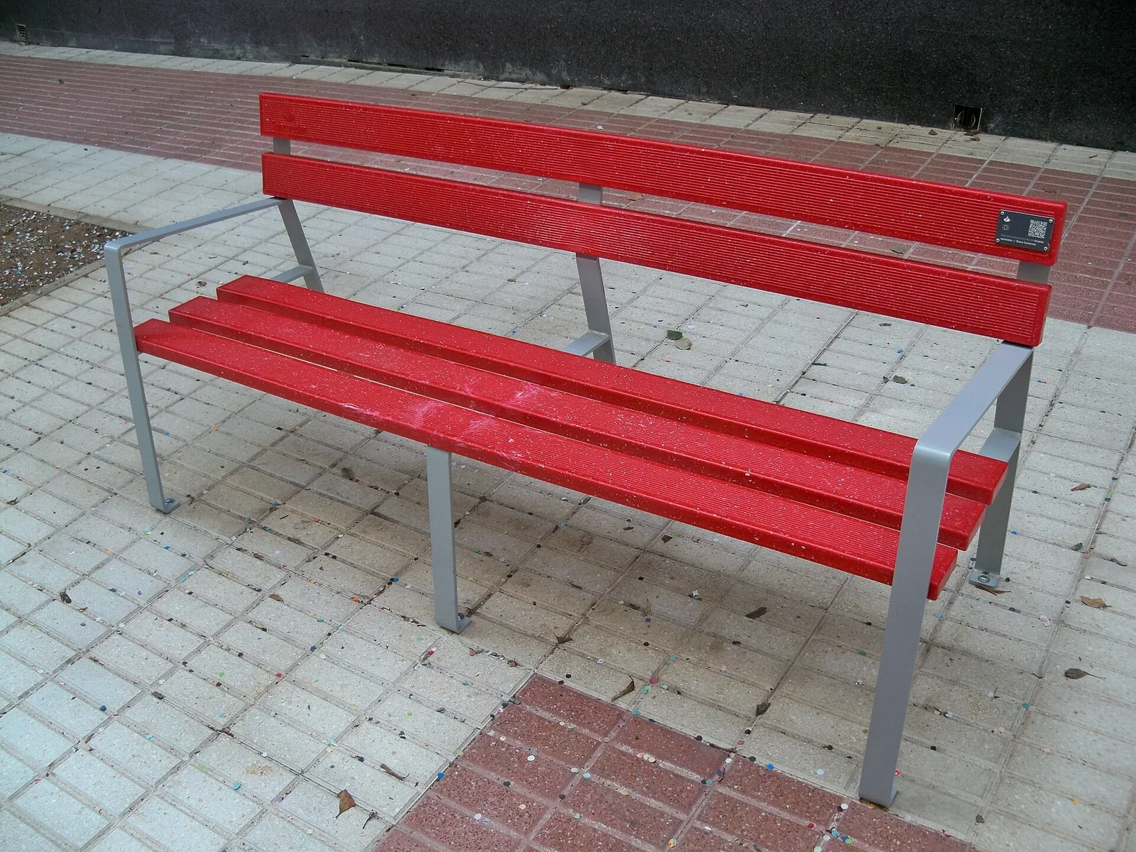 Red recycled-plastic bench with metal legs on a paved street in Málaga