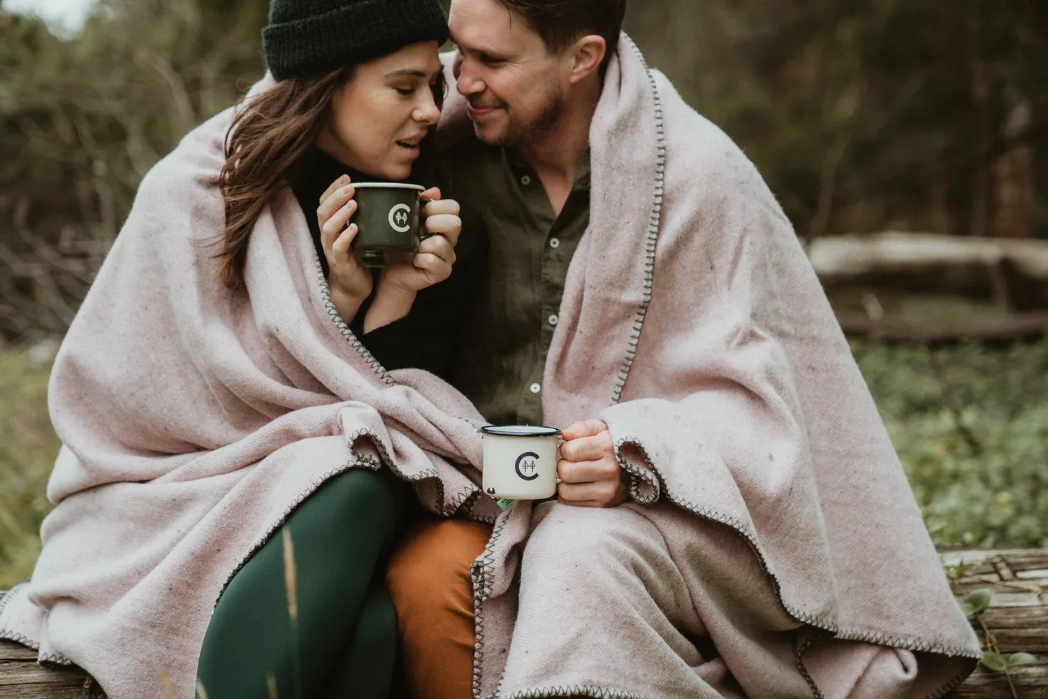 Two people sharing a pale blanket and holding cups outdoors.